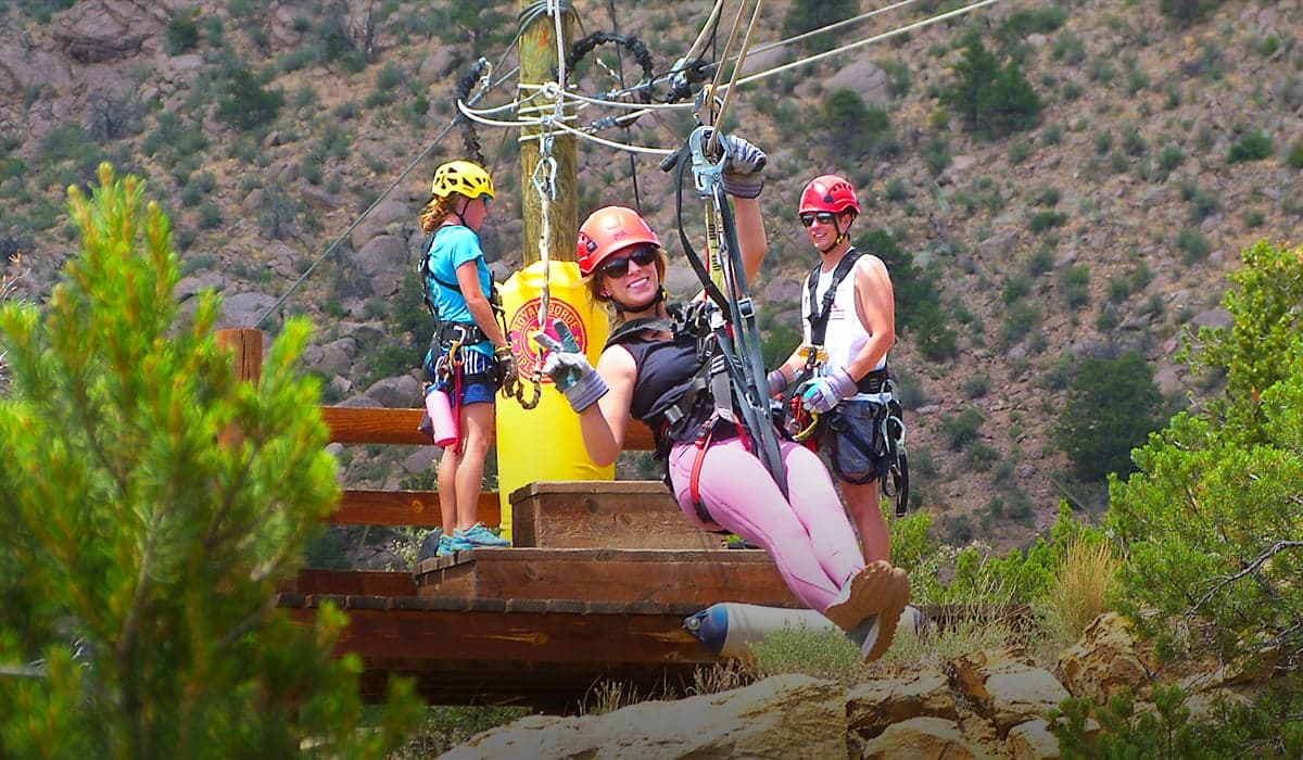 A smiling rider in a helmet and harness pushing off a wooden zipline platform, with two guides behind her