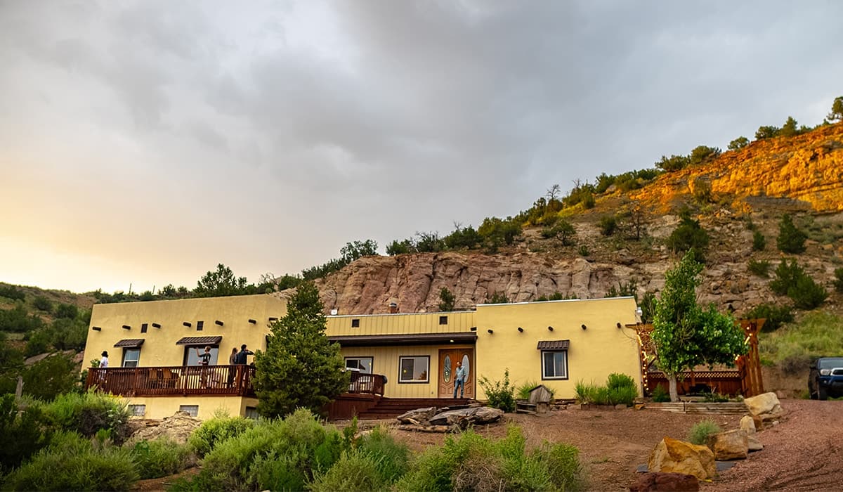 A stucco ranch house with a long wooden deck below a red rock hillside at dusk
